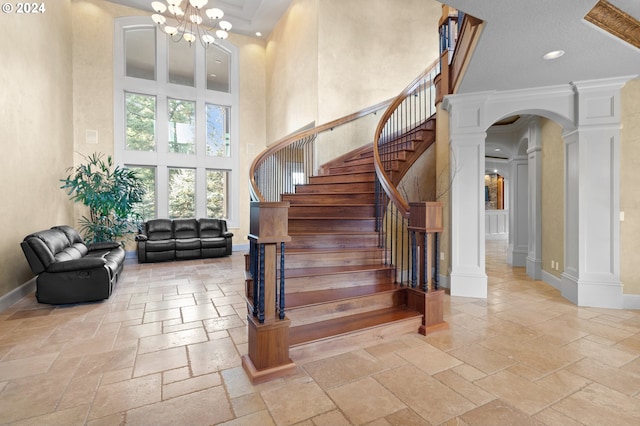 staircase featuring crown molding, an inviting chandelier, a towering ceiling, light tile floors, and ornate columns