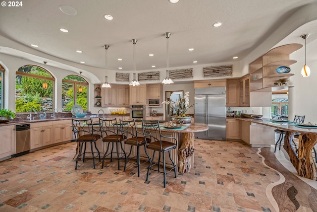 kitchen featuring built in appliances, decorative light fixtures, a kitchen island with sink, and light brown cabinetry