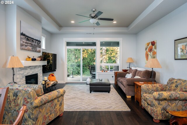 living room with ceiling fan, dark wood-type flooring, a stone fireplace, and a tray ceiling