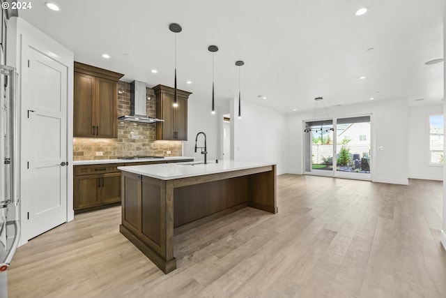 kitchen featuring an island with sink, pendant lighting, sink, light hardwood / wood-style flooring, and wall chimney range hood