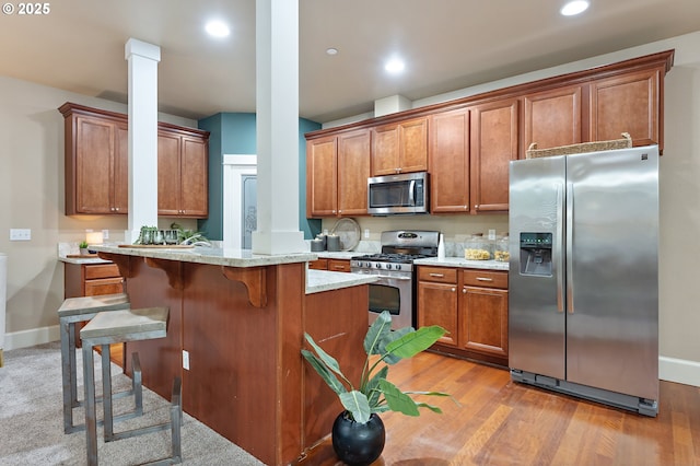 kitchen with light stone countertops, light wood-type flooring, appliances with stainless steel finishes, a breakfast bar area, and decorative columns