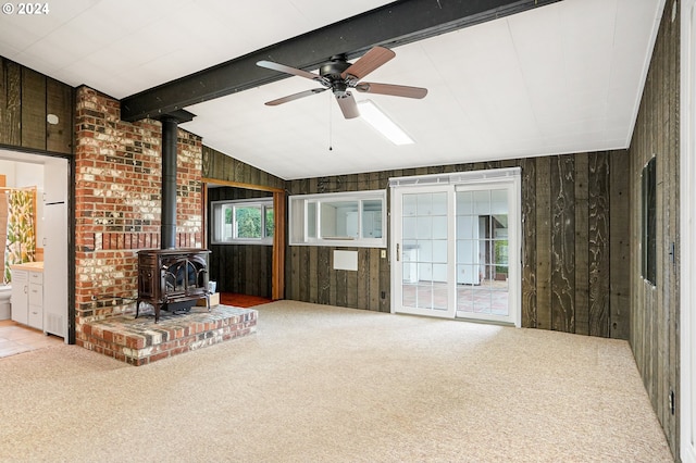 unfurnished living room featuring a wood stove, ceiling fan, lofted ceiling with beams, wood walls, and light colored carpet