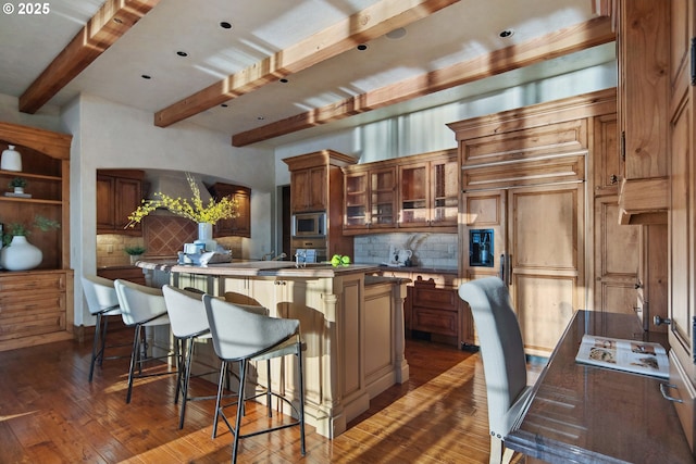 kitchen featuring tasteful backsplash, stainless steel microwave, dark hardwood / wood-style floors, an island with sink, and beam ceiling