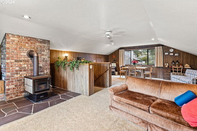 carpeted living room with vaulted ceiling, wood walls, a wood stove, a textured ceiling, and ceiling fan