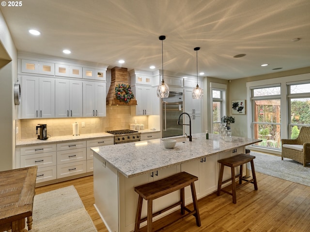 kitchen with custom exhaust hood, high quality appliances, an island with sink, decorative light fixtures, and white cabinetry