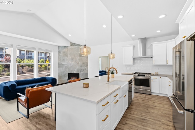 kitchen with wall chimney exhaust hood, an island with sink, hanging light fixtures, stainless steel appliances, and white cabinetry