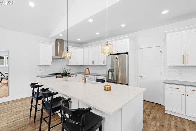 kitchen featuring wall chimney range hood, appliances with stainless steel finishes, light wood-type flooring, pendant lighting, and a center island with sink