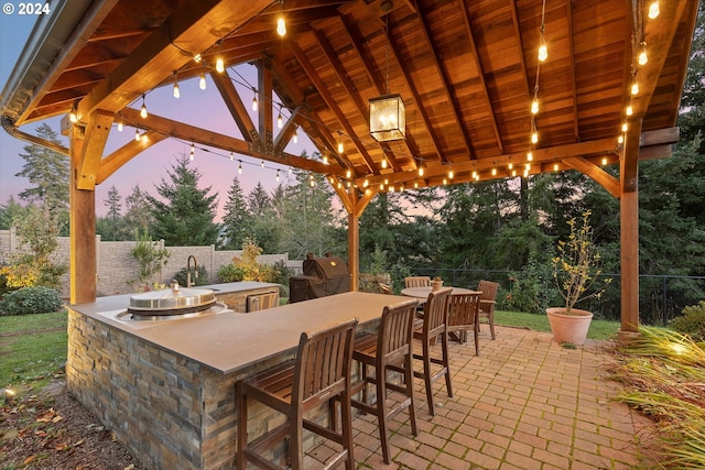 patio terrace at dusk with a bar, area for grilling, an outdoor kitchen, and a gazebo