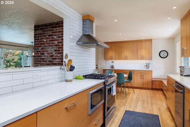 kitchen featuring wall chimney range hood, light countertops, appliances with stainless steel finishes, light wood-style flooring, and decorative backsplash