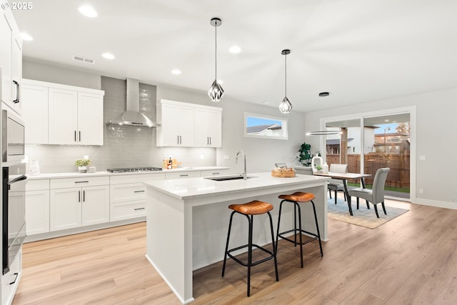 kitchen with white cabinetry, a center island with sink, hanging light fixtures, wall chimney range hood, and sink