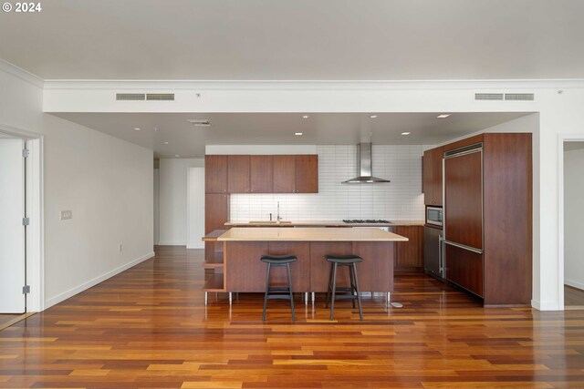 kitchen featuring wall chimney range hood, appliances with stainless steel finishes, backsplash, a kitchen breakfast bar, and a center island