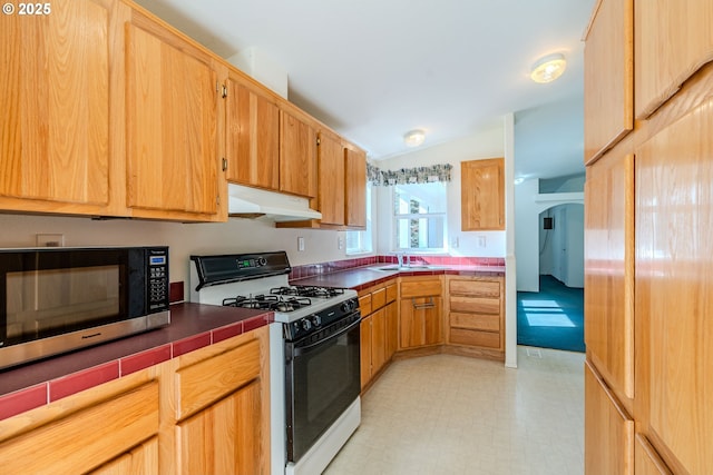 kitchen with tile countertops, under cabinet range hood, gas range oven, light floors, and stainless steel microwave