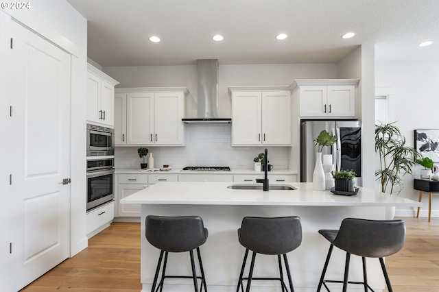 kitchen featuring a center island with sink, light hardwood / wood-style flooring, wall chimney exhaust hood, appliances with stainless steel finishes, and white cabinetry