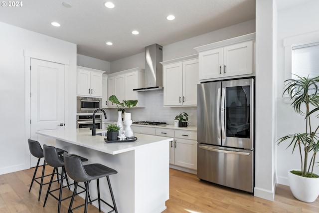 kitchen with light wood-type flooring, wall chimney exhaust hood, stainless steel appliances, a kitchen island with sink, and white cabinetry