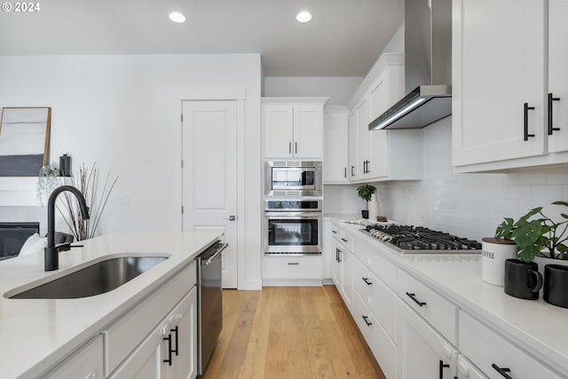 kitchen with white cabinets, wall chimney range hood, sink, appliances with stainless steel finishes, and light hardwood / wood-style floors