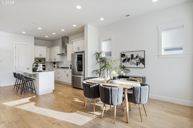 kitchen featuring appliances with stainless steel finishes, a kitchen island with sink, wall chimney range hood, white cabinetry, and a breakfast bar area