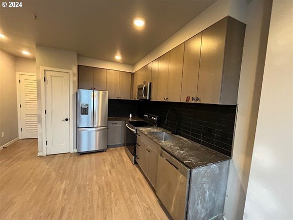 kitchen with appliances with stainless steel finishes, light wood-type flooring, tasteful backsplash, sink, and dark stone countertops