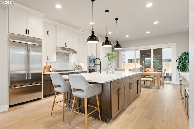 kitchen with pendant lighting, a kitchen island with sink, white cabinets, light hardwood / wood-style flooring, and stainless steel appliances