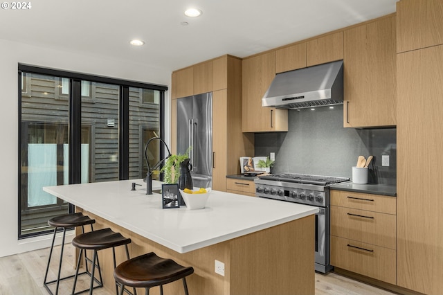 kitchen with wall chimney range hood, light hardwood / wood-style floors, high end appliances, and a kitchen island with sink