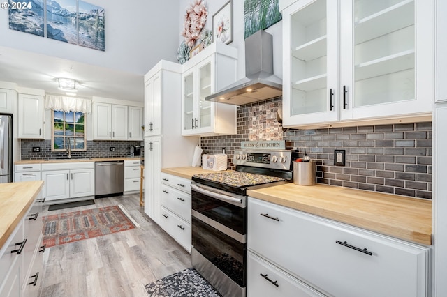 kitchen featuring wall chimney range hood, decorative backsplash, wooden counters, white cabinets, and appliances with stainless steel finishes