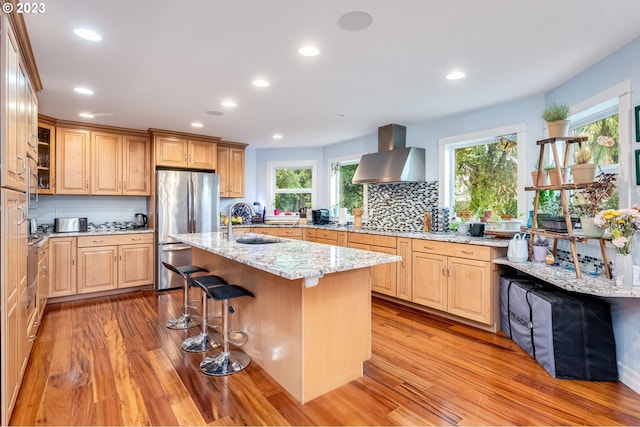 kitchen with light stone countertops, light wood-type flooring, a kitchen island with sink, exhaust hood, and stainless steel refrigerator