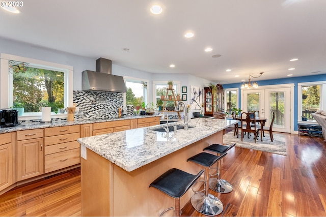 kitchen with sink, wall chimney range hood, dark hardwood / wood-style flooring, an island with sink, and light brown cabinetry