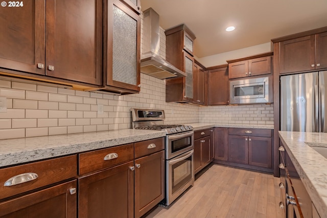 kitchen featuring light stone countertops, wall chimney range hood, tasteful backsplash, appliances with stainless steel finishes, and light wood-type flooring