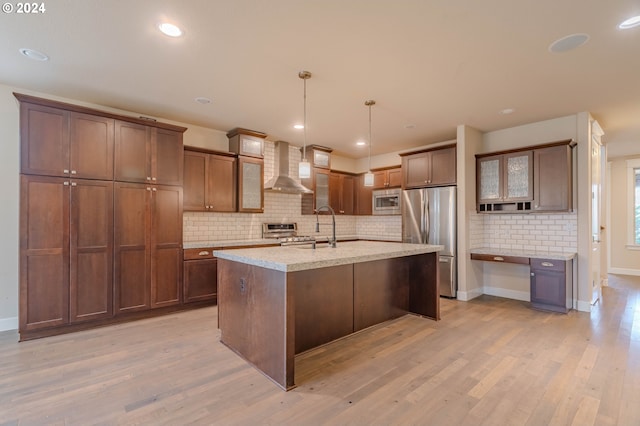 kitchen with light hardwood / wood-style flooring, wall chimney exhaust hood, an island with sink, decorative light fixtures, and stainless steel appliances