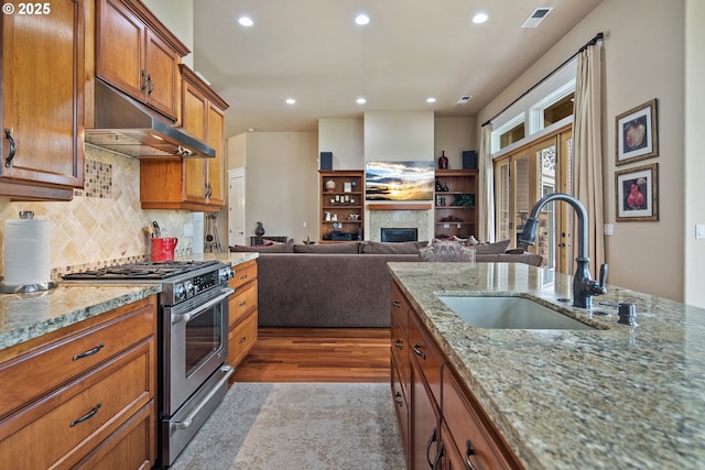 kitchen with a glass covered fireplace, brown cabinets, stainless steel stove, under cabinet range hood, and a sink