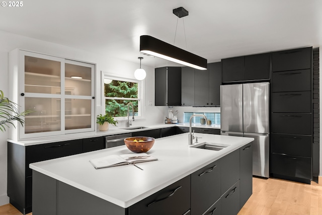 kitchen with a kitchen island with sink, appliances with stainless steel finishes, light wood-type flooring, pendant lighting, and sink