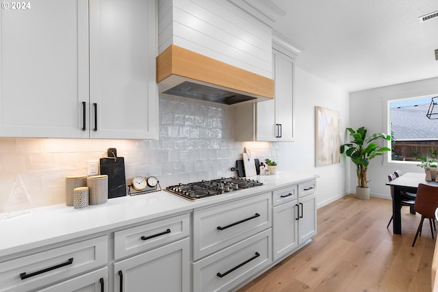 kitchen featuring stainless steel gas stovetop, white cabinets, decorative backsplash, custom exhaust hood, and light hardwood / wood-style flooring