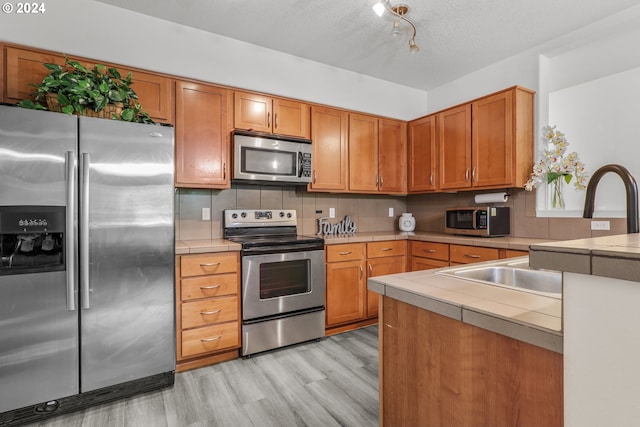 kitchen with tile counters, decorative backsplash, brown cabinetry, light wood-style flooring, and appliances with stainless steel finishes