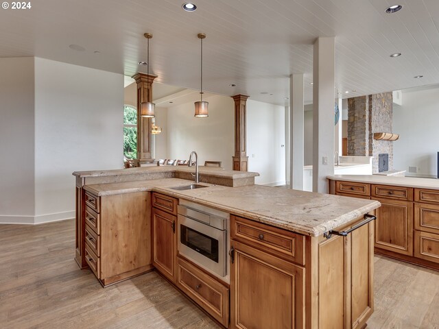 kitchen with decorative light fixtures, an island with sink, sink, stainless steel microwave, and light hardwood / wood-style floors