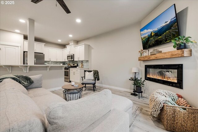 living room with baseboards, a ceiling fan, a glass covered fireplace, light wood-type flooring, and recessed lighting