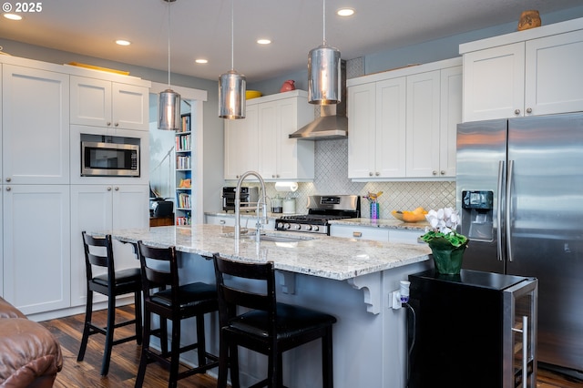 kitchen featuring decorative light fixtures, white cabinetry, stainless steel appliances, a center island with sink, and wall chimney exhaust hood