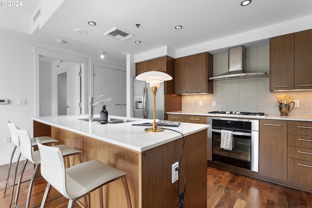 kitchen featuring appliances with stainless steel finishes, sink, an island with sink, dark hardwood / wood-style flooring, and wall chimney exhaust hood