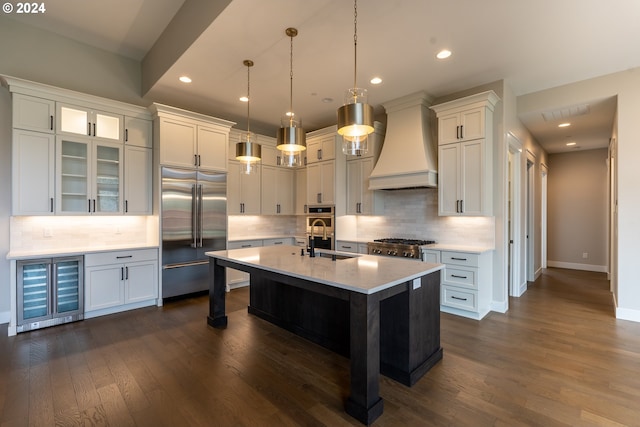 kitchen featuring premium range hood, stainless steel appliances, a kitchen island with sink, dark wood-type flooring, and decorative light fixtures