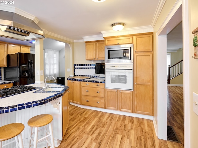 kitchen with tile counters, range hood, light wood-type flooring, black appliances, and ornamental molding