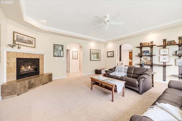 living room with a tiled fireplace, ornamental molding, light colored carpet, ceiling fan, and a tray ceiling