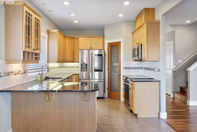 kitchen with light brown cabinets, stainless steel appliances, a peninsula, a sink, and dark stone countertops