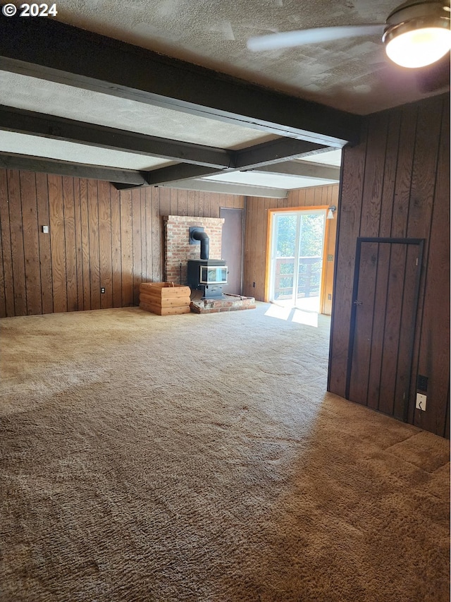 unfurnished living room featuring carpet, wood walls, beamed ceiling, and a wood stove