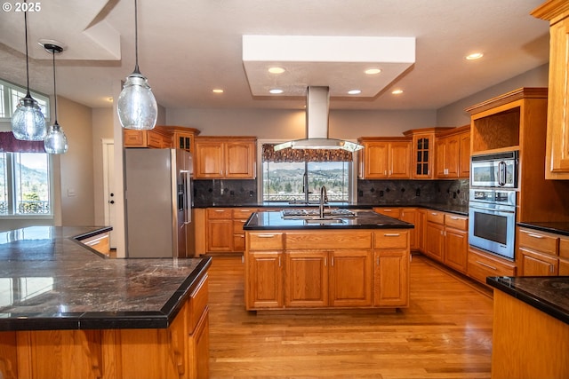 kitchen featuring brown cabinets, an island with sink, stainless steel appliances, island range hood, and glass insert cabinets