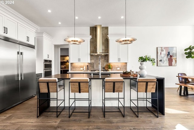 kitchen featuring dark wood-type flooring, wall chimney range hood, appliances with stainless steel finishes, and a kitchen island with sink