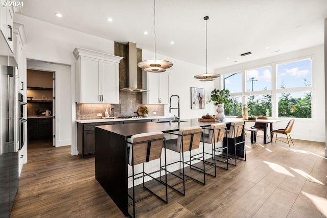 kitchen featuring decorative light fixtures, dark hardwood / wood-style flooring, tasteful backsplash, a center island with sink, and wall chimney exhaust hood