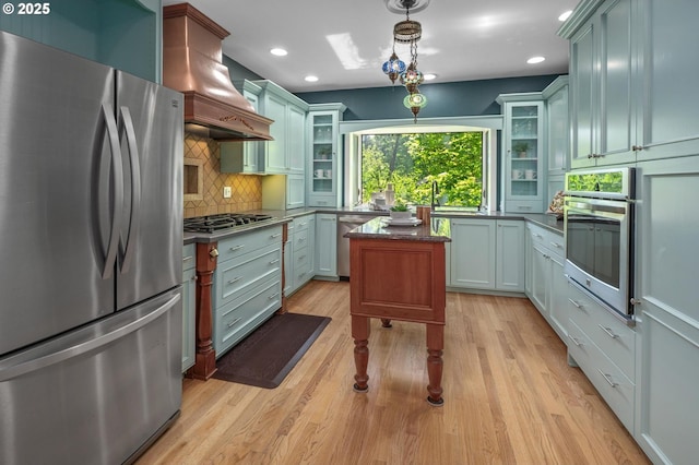 kitchen featuring backsplash, premium range hood, glass insert cabinets, stainless steel appliances, and light wood-style flooring