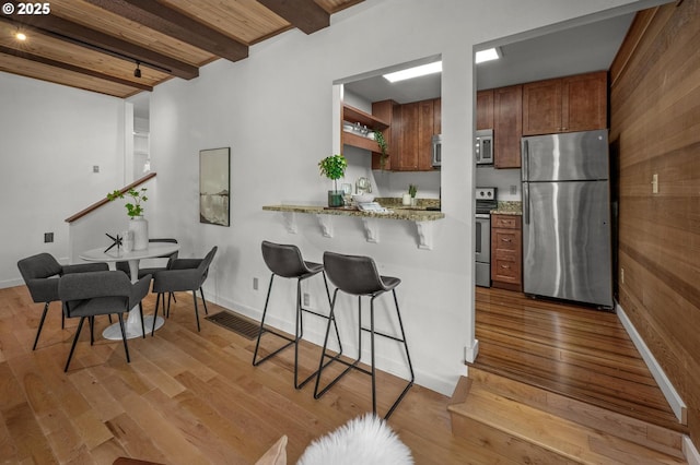 kitchen featuring beam ceiling, a kitchen breakfast bar, brown cabinetry, stainless steel appliances, and wood finished floors