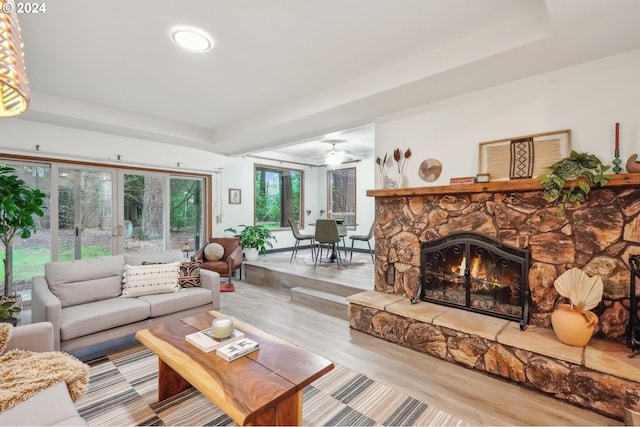 living room with a raised ceiling, ceiling fan, a stone fireplace, and light hardwood / wood-style flooring