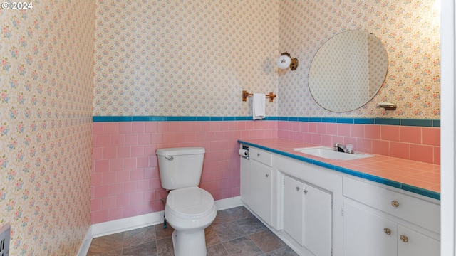 bathroom with toilet, wallpapered walls, tile walls, wainscoting, and vanity