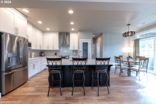 kitchen with wall chimney exhaust hood, stainless steel appliances, pendant lighting, a kitchen island with sink, and white cabinets