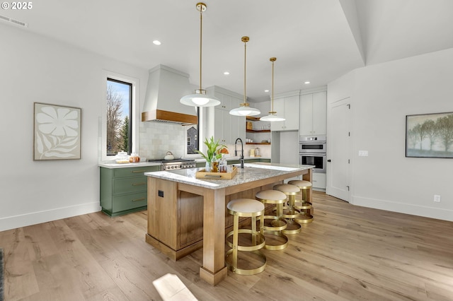 kitchen featuring decorative light fixtures, a center island with sink, tasteful backsplash, white cabinetry, and premium range hood